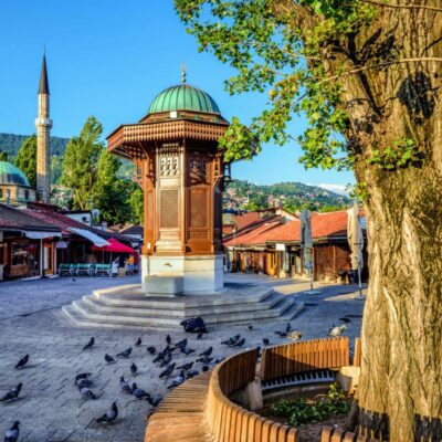 Public square with a carved wooden gazebo featuring a green dome, pigeons on the stone pavement, and a large tree in the foreground.