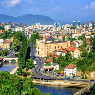 Panoramic cityscape with a river, bridge, and colorful buildings against green hills and a blue sky.