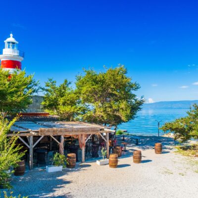 Red-and-white lighthouse peeks through green trees by a lakeside, with a rustic outdoor seating area in the foreground and clear blue sky above.
