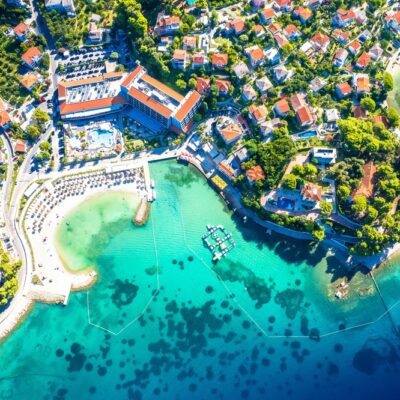 Aerial view of a seaside town with a turquoise bay, sandy beach, and red-roofed houses surrounded by green trees.