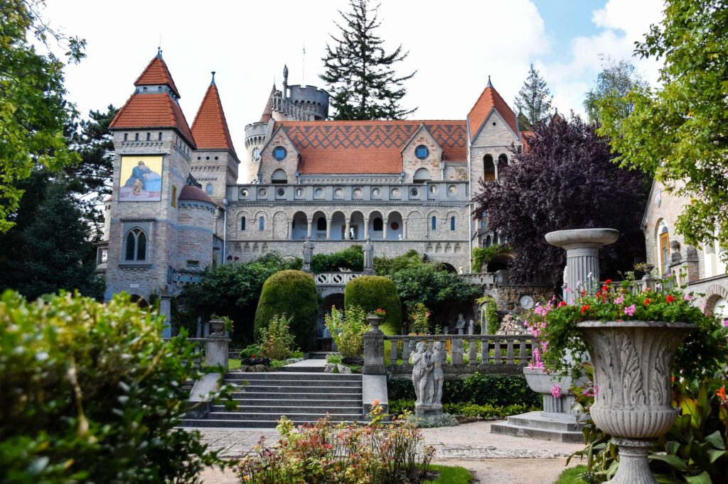 Castle-like stone building with red tiled roof and multiple towers, set in a formal garden with statues, arches, and a stone urn in the foreground.